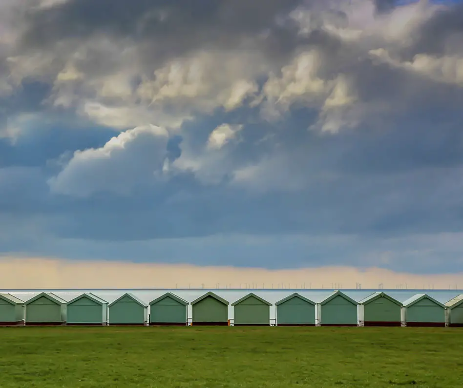 A row of identical green beach huts between a lawn and the sea, heavy clouds above.