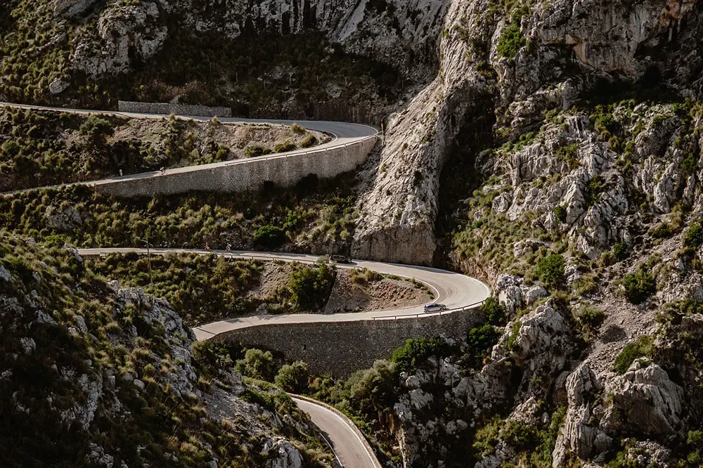 A wildly snaking mountain road cut into a rocky terrain peppered with green bushes.