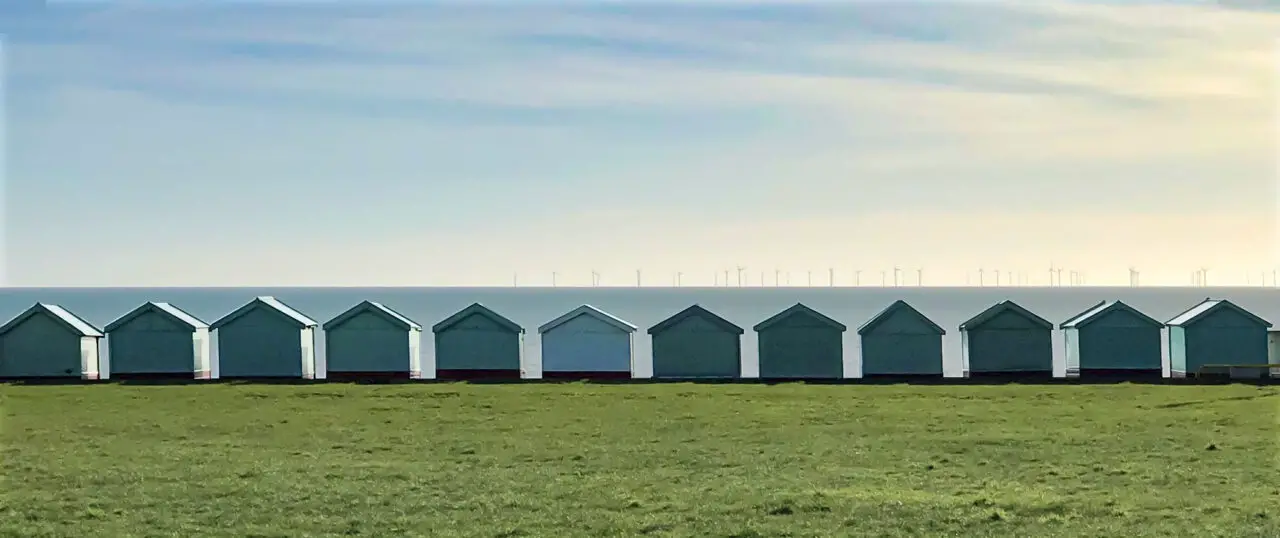 A uniform row of green huts between the sea and a lawn.